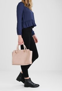 Borsa strutturata rosa chiaro con doppi manici superiori, texture liscia e cuciture sottili; abbinata a jeans skinny neri e stivaletti.