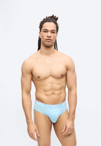 Μυώδης άνδρας με μακριά κοτσίδα dreadlocks που φοράει γαλάζια εσώρουχα, στέκεται μπροστά σε απλό ανοιχτό γκρι φόντο, κοιτάζοντας μπροστά.