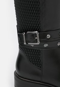 Botte en cuir noir avec tige tissée texturée, boucle latérale et détails décoratifs en métal le long de la sangle. Talon épais.