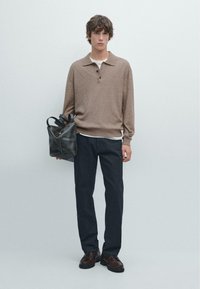 Giovane uomo che indossa un maglione marrone, pantaloni scuri e scarpe scure, mentre tiene una borsa di pelle nera contro uno sfondo grigio semplice.