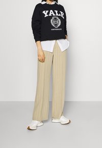 Felpa nera con grafica "Yale University", camicia bianca con colletto sotto, pantaloni larghi a righe beige e sneakers bianche con suola marrone.