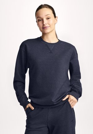 Kobieta o ciemnych włosach związanych do tyłu, ubrana w ciemnoniebieską bluzę i pasujące spodnie, stojąca z rękami w kieszeniach, na neutralnym tle.