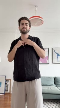 Camicia nera a maniche corte con colletto e tessuto a trama, abbinata a pantaloni chiari con pieghe.