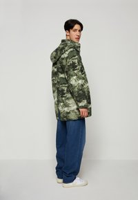 Zöld, álcázó mintás dzseki, kapucnival, texturált anyagból készült. Hosszú, laza szabású kék farmernadrággal és fehér sportcipőkkel viselve.