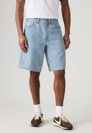 Homme portant un short en jean bleu clair avec un écusson du blason des États-Unis, des chaussettes blanches, des baskets marron et blanches, et un t-shirt blanc uni, debout devant un fond gris.