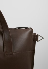Coin d'un sac cabas en cuir marron avec une poignée, une fermeture éclair sur le dessus partiellement visible, et un anneau métallique triangulaire sur le côté sur un fond uni.