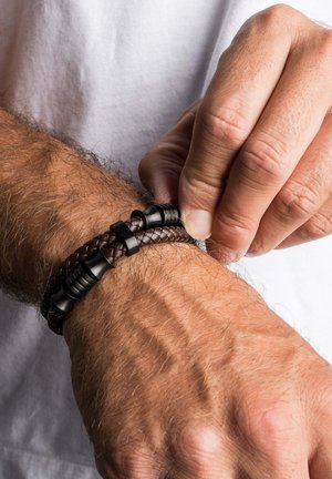 Man fastening a brown braided leather bracelet with black magnetic clasp on hairy wrist, wearing white shirt.