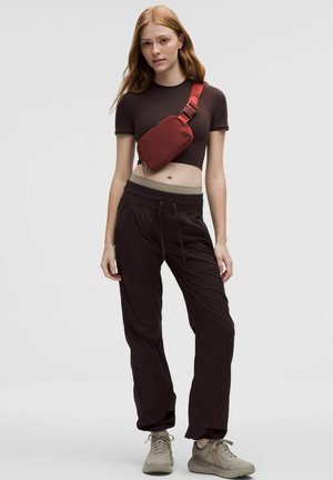 Geantă crossbody roșie, top scurt maro, pantaloni joggers negri și adidași bej; design elegant cu curea ajustabilă, talie elastică și buzunare.