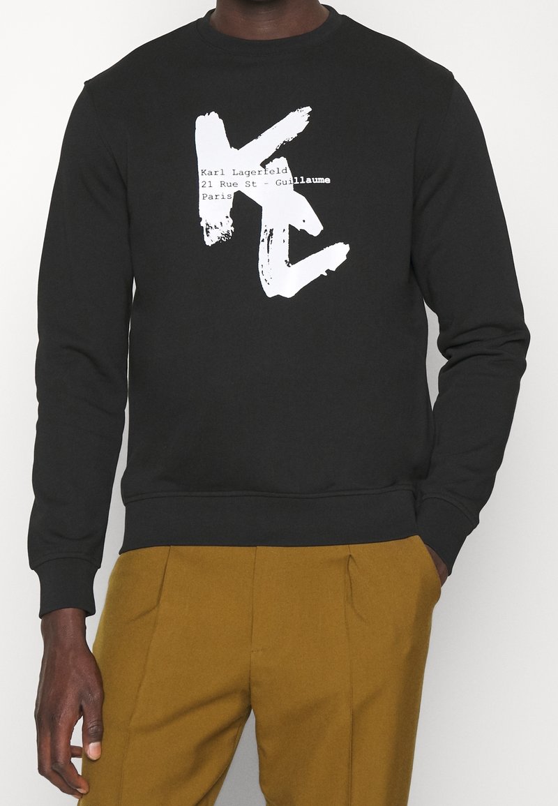Chemise noire à manches longues avec de grandes lettres blanches en traits de pinceau "KL" et texte incluant "Karl Lagerfeld" et "Paris,", associée à un pantalon moutarde.