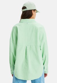 Kvinna i ljusgrön corduroyskjorta, matchande grön keps och blå jeans, sedd bakifrån mot en vit bakgrund.
