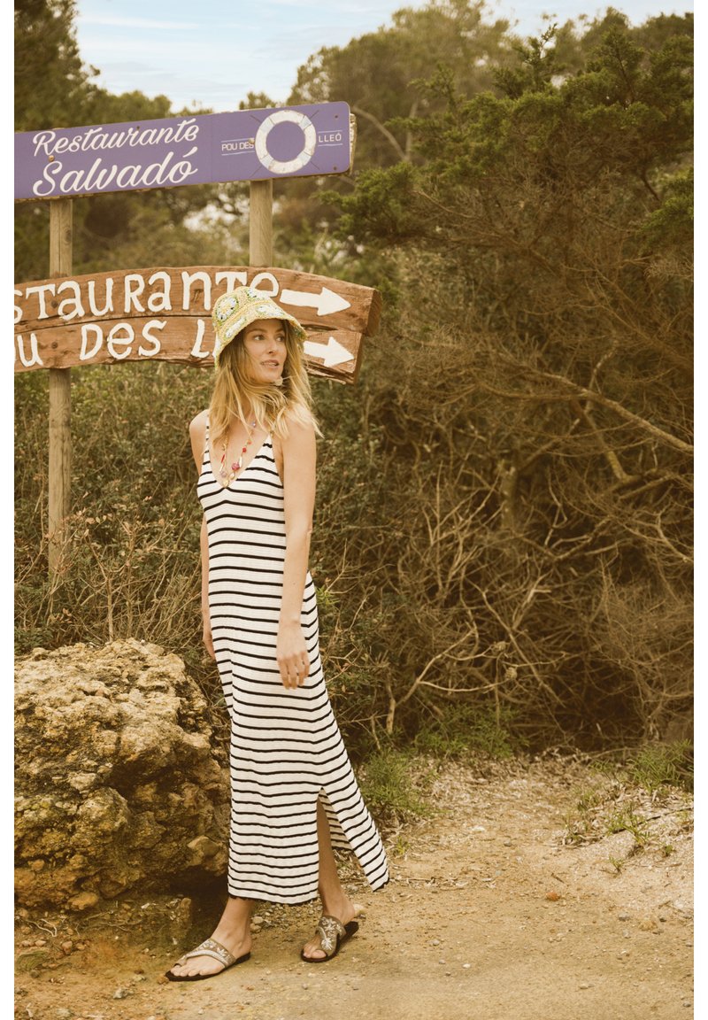 Abito maxi a righe senza maniche con spacco laterale, indossato con sandali decorati; accessorato con un cappello colorato e a fantasia. Lo sfondo include segnali in legno.