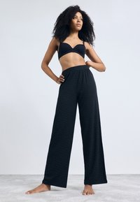 Svartmønstret vide bukser med elastisk midjebånd, kombinert med en svart bh, båret av en modell med langt, krøllete hår.