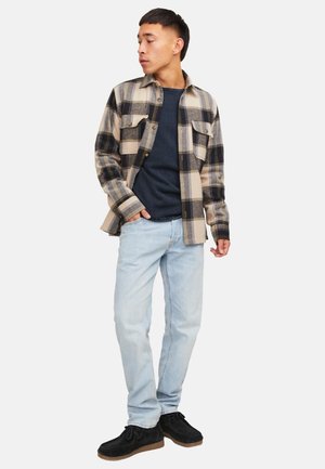 Ung mand iført en beige og sort ternet skjorte over en mørkeblå sweater, lyseblå jeans og sorte afslappede sko, stående og kigger sidelæns.