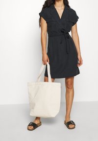 Abito nero a maniche corte con vita alta, dettagli a bottoni e scollatura a V. Abbinato a una borsa a tracolla in tela color crema e sandali neri.