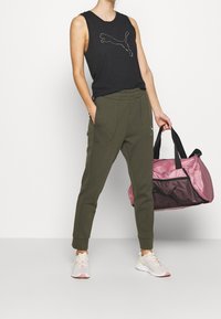 Schwarzes ärmelloses Oberteil mit einem weißen Logo, olivgrüne Sweatpants und eine pink/schwarze Sporttasche, kombiniert mit weißen Sneakers mit pinken Akzenten.