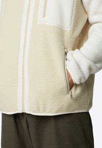 Lys beige fleecejakke med glidelåslommer, med et mykt teksturert stoff og kontrasterende hvite innsatser langs fronten.