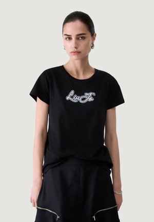 Mujer con cabello oscuro, con una camiseta negra con texto de logo blanco y falda negra a juego con dos cremalleras delanteras, de pie sobre un fondo liso.