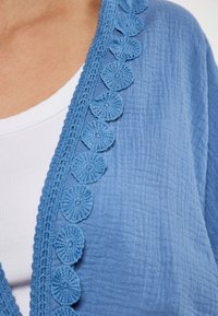 Cardigan de culoare albastru deschis, realizat dintr-un material texturat, având o dantelă croșetată cu margini ondulate pe partea din față deschisă și o căptușeală albă vizibilă dedesubt.