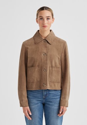 Frau trägt eine hellbraune Veloursleder-Jacke mit großen Fronttaschen und blauen Jeans, steht vor einem schlichten weißen Hintergrund.