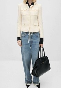 Cremefarbener Tweed-Blazer mit zwei Fronttaschen, kombiniert mit lockeren blauen Jeans und einer schwarzen Handtasche. Spitz zulaufende schwarze Pumps vervollständigen das Outfit.