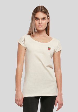 Jeune femme portant un t-shirt à manches courtes blanc cassé avec un petit motif fraise à gauche sur la poitrine et un pantalon noir.