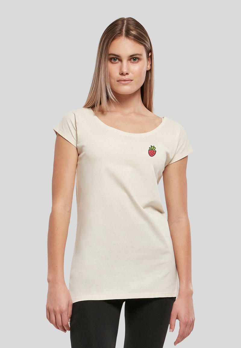 Jeune femme portant un t-shirt à manches courtes blanc cassé avec un petit motif fraise à gauche sur la poitrine et un pantalon noir.