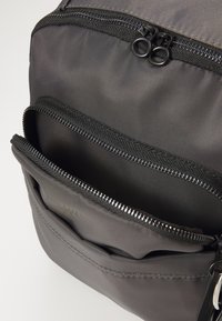Šedý látkový batoh s několika zipovými přihrádkami, s hladkými texturami a jemnými detaily šití. Zipy mají černý kovový rám.