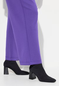 Lila weit geschnittene Hosen und schwarze Sockenabsatzschuhe mit quadratischen Absätzen und spitzen Zehen, die eine glatte Textur und ein minimalistisches Design aufweisen.