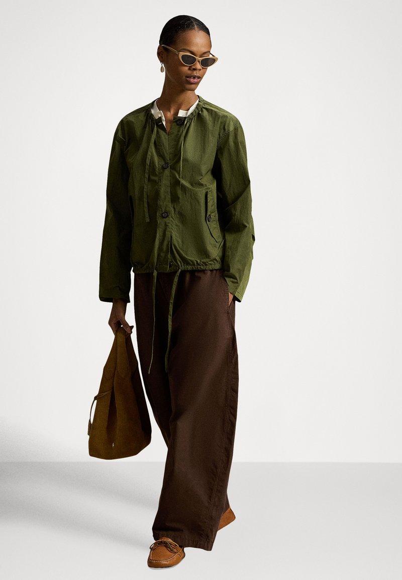 Woman wearing olive green jacket, wide brown pants, tan shoes, and cat-eye sunglasses, holding a brown suede bag, standing on a plain background.