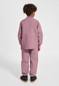 Dziecko z kręconymi włosami stoi tyłem, ubrane w pikowaną, zakurzoną różową kurtkę i pasujące spodnie oraz czarne buty na białym tle.