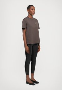 Brun bomull t-skjorte med korte ermer og en liten logo, kombinert med sorte skinnleggins og flate svarte sko.