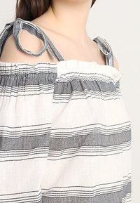 Off-shoulder topp i lätt tyg med horisontella svartvita ränder, med knutna axel detaljer och rynkad krage.