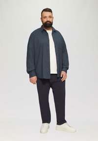 Blauw-wit geruite knoopshirt over een wit T-shirt, gecombineerd met donkere broeken en witte sneakers. Eenvoudige achtergrond.