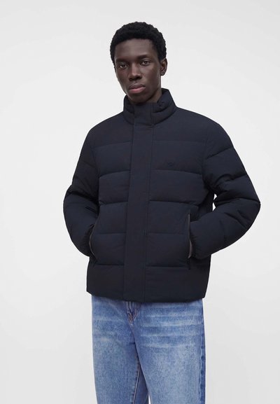 Homme portant une doudoune noire les mains dans les poches et un jean bleu, debout devant un fond clair uni.
