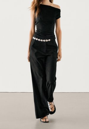 Femme portant un top noir épaules dénudées, un pantalon noir à jambes larges avec une ceinture circulaire blanche, et des sandales noires à brides marchant vers l'avant.