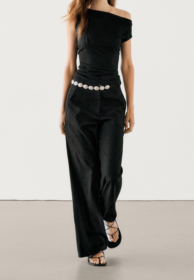 Femme portant un top noir épaules dénudées, un pantalon noir à jambes larges avec une ceinture circulaire blanche, et des sandales noires à brides marchant vers l'avant.