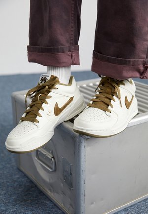 Białe i brązowe sneakersy Nike z sznurowadłami, noszone z białymi skarpetkami i podwiniętymi brązowymi spodniami, stopy spoczywające na metalowym pudełku.