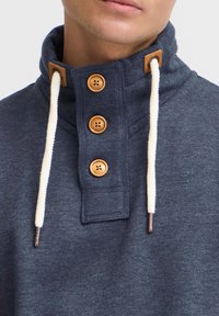 Jersey azul marino con cuello alto, que presenta botones de madera y un diseño tipo toggle. Confeccionado con una tela suave y acabado texturizado, y cordones blancos en contraste.
