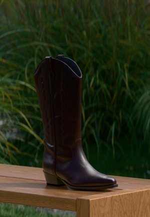 Bota de cowboy de cuero marrón oscuro con tacón bajo, de pie sobre un banco de madera al aire libre con hierba alta y verde de fondo.