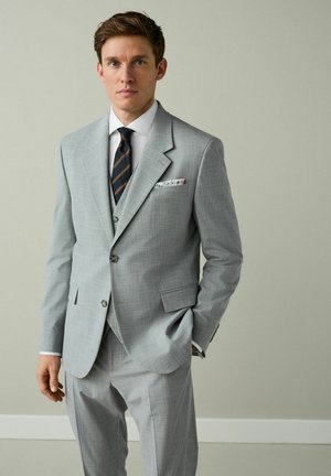 Młody mężczyzna ubrany w jasnoszary garnitur trzyczęściowy, białą koszulę, granatowy krawat w paski oraz kwiatową poszetkę, stojący z jedną ręką w kieszeni.