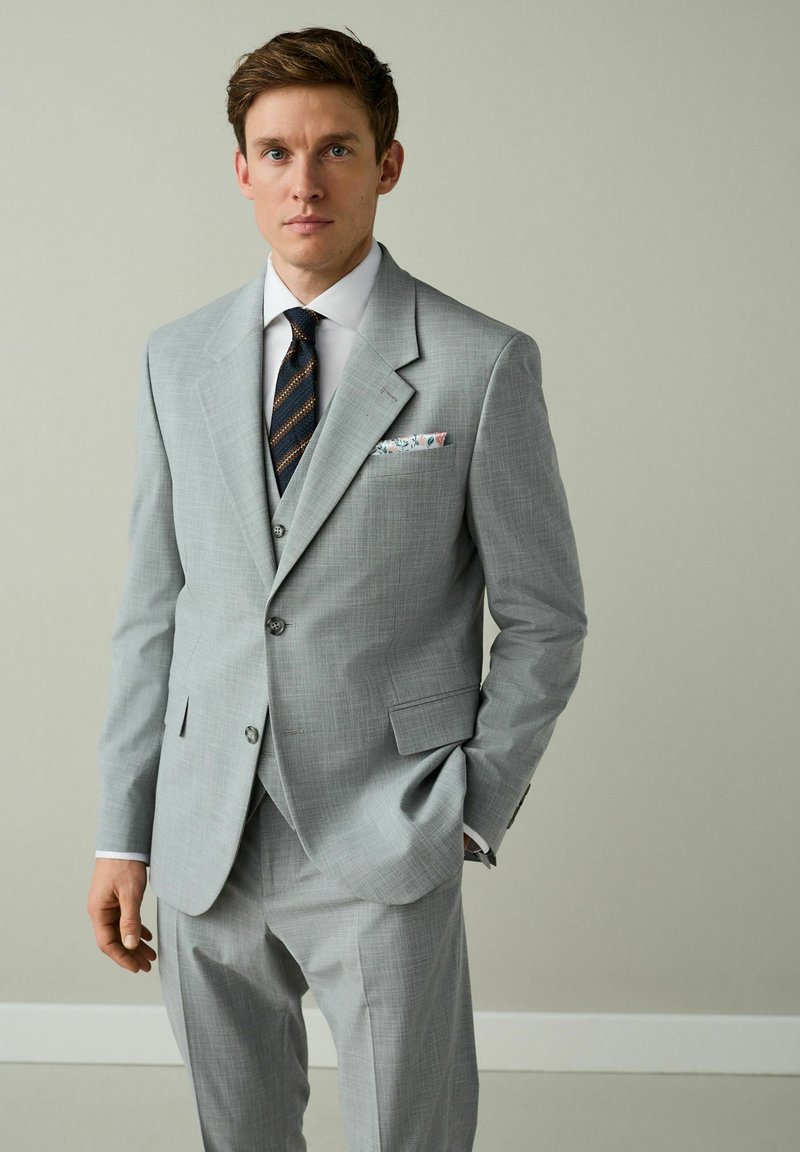 Junger Mann trägt einen hellgrauen dreiteiligen Anzug, weißes Hemd, gestreifte marineblaue Krawatte und ein florales Einstecktuch, steht mit einer Hand in der Tasche.