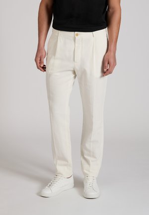 Hombre con pantalones a medida color off-white con pliegues, zapatillas blancas y camisa negra, de pie sobre un fondo neutro.