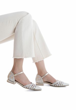 Zapatos planos con correa en el tobillo en color plateado metálico con diseño tejido, punta estrecha y tacón bajo, combinados con vaqueros blancos recortados.