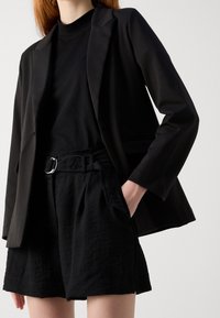 Blazer noir et short noir assorti. Le blazer a une coupe ajustée avec un seul bouton ; le short est doté d'une ceinture avec une boucle argentée et de plis.