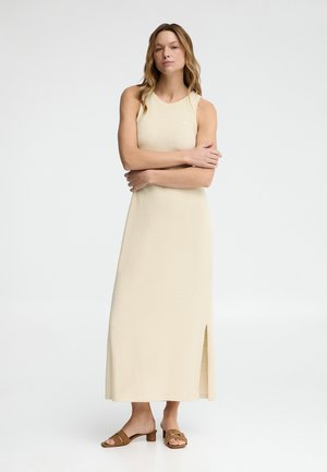 Frau mit langen Haaren, die ein ärmelloses cremefarbenes Maxikleid mit Seitenschlitz und braunen Sandalen mit Absatz trägt, steht mit verschränkten Armen.