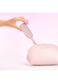 Hånd med rosa negler som holder en rosa luftet hårbørste over en matchende rosa teksturert pose med "brushworks"-logo.