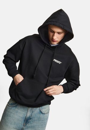 Młoda osoba ubrana w czarną bluzę z kapturem z logo "PROSTO", ręce w przedniej kieszeni i jasnoniebieskie dżinsy na jednolitym tle.