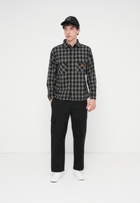 Chemise en flanelle à carreaux noirs et gris avec deux poches avant, associée à un pantalon cargo noir et des baskets blanches. La casquette comporte un écusson logo.