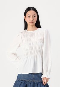 Hvit langermet bluse med smock-struktur, rund halslinning og samlet midje, kombinert med en denskjørt med lagdelt design.