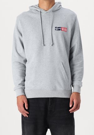 Homme portant un sweat à capuche gris clair avec poche avant et logo Tommy Jeans sur la poitrine, associé à un pantalon noir, debout devant un fond uni.